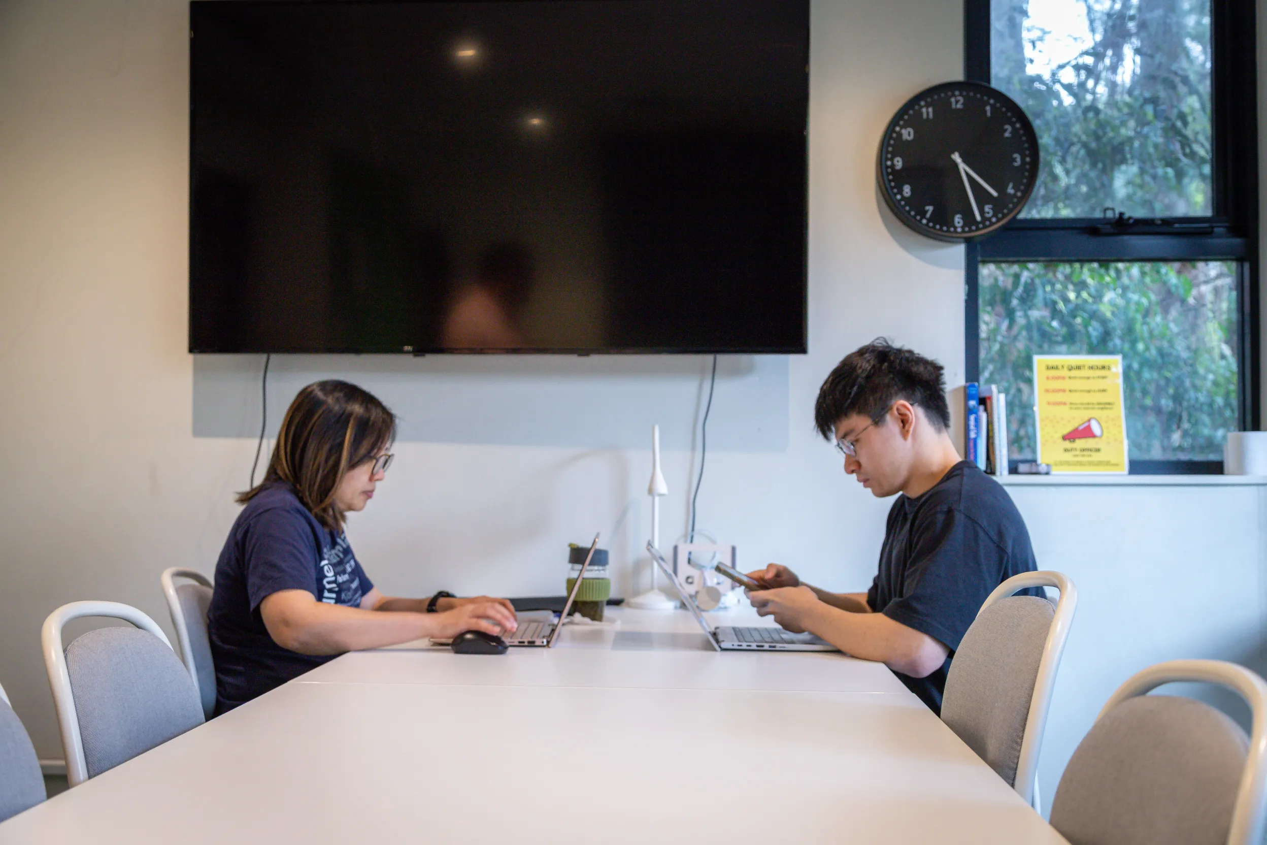 Two residents studying on laptops in a study room at Robert Menzies College, with glass doors opening onto a leafy deck.