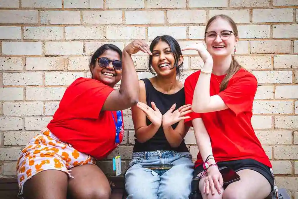 Three smiling students sit against a brick wall, two in red college t-shirts, together making heart shapes with their hands.