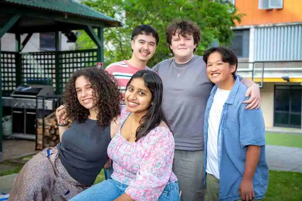 Five students smile together with arms around each other in a leafy college courtyard, with a gazebo and barbecue behind them.
