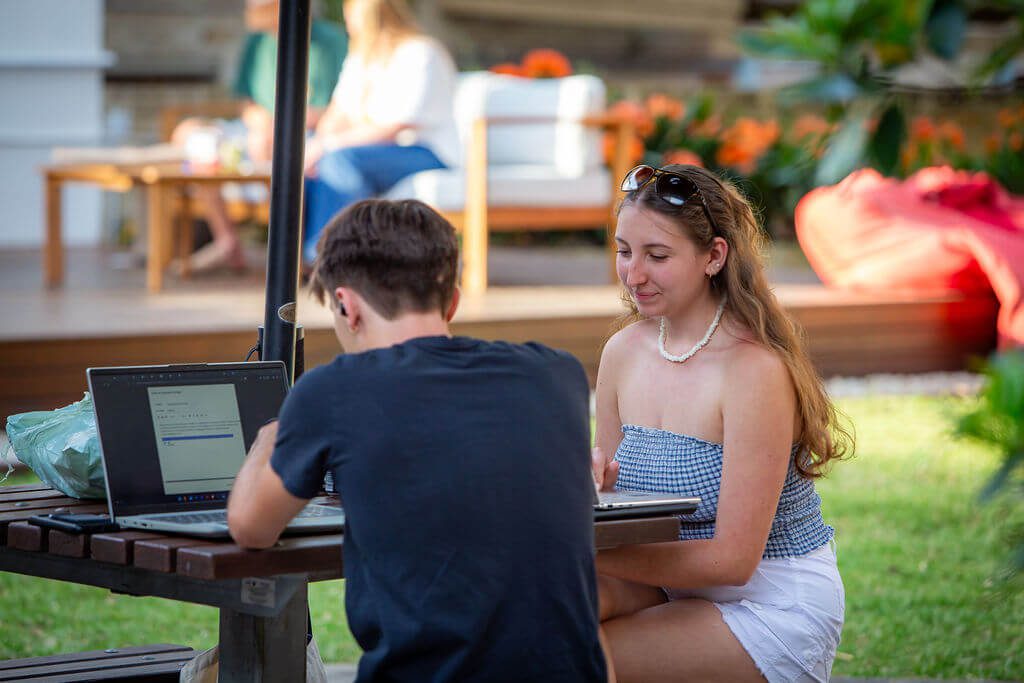 Two residents studying on laptops at an outdoor table on the lawn at Robert Menzies College, with others relaxing on a deck behind.